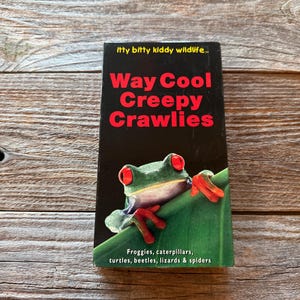 May include: A black book titled "Way Cool Creepy Crawlies" with a green frog illustration. The book also lists "Froggies, caterpillars, turtles, beetles, lizards & spiders". The book is on a wooden surface.
