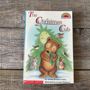 Puede incluir: Un libro infantil titulado "The Christmas Cub" con una ilustración de un oso pardo, una ardilla y un conejo. La portada presenta un árbol de Navidad con una cara sonriente y pequeños pájaros. El libro es de Justine Korman Fontes e ilustrado por Lucinda McQueen.