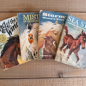 Puede incluir: Cuatro libros infantiles antiguos con ilustraciones de caballos. Los títulos incluyen "King of the Wind", "Mist of Chincoteague", "Storm, Misty's Foal" y "Sea Star, Orphan of Chincoteague". Los libros tienen cubiertas coloridas y están sobre una superficie de madera.