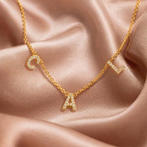 May include: A gold-toned chain necklace featuring three individual letter charms spelling CAL. Each letter is encrusted with small, clear, sparkling stones. The delicate metal chain connects the letters, creating a spaced-out, dangling letter design.