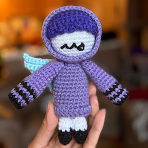 May include: A purple crocheted doll with a white face and black eyes. The doll is wearing a purple hooded sweater with black and white stripes on the sleeves. The doll has a light blue wing on its back.