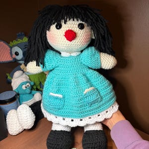 May include: A crocheted doll with black hair, a red nose, and a blue dress. The doll is wearing black shoes and has a white crocheted collar.