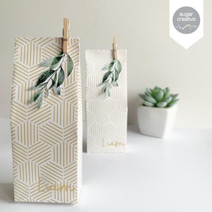 May include: Two white gift bags with gold geometric patterns and green leafy embellishments. The bags are tied with wooden clothespins and have the name "Liam" written on them.
