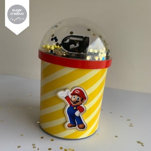 Może przedstawiać: Żółty i biały, pasiasty pojemnik cylindryczny z przezroczystą kopułą na górze. Pojemnik ma czerwoną taśmę wokół górnej części i naklejkę z postacią z gry wideo Mario. Kopuła zawiera czarny i szary obiekt w kształcie pocisku oraz złoty brokat.