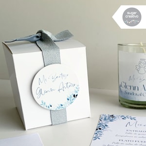 Puede incluir: Caja de regalo blanca con una cinta azul y una pegatina redonda que dice "Mi Bautizo Glenn Arturo" en letra azul con un diseño floral de acuarela. La caja está al lado de una vela de vidrio transparente con una etiqueta azul y blanca que dice "Mi Bautizo Glenn A Sandoval C".