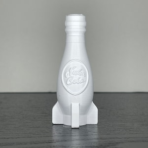 May include: A white 3D-printed bottle shaped like a rocket, featuring the "Nuka Cola" logo. The bottle has a textured surface and a rocket-like base. The design is a nod to the Fallout video game series.