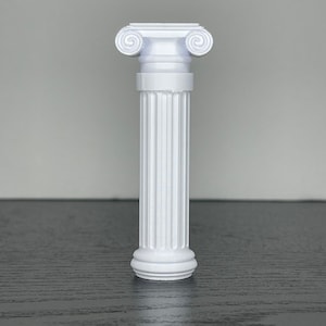 May include: A white, miniature Ionic column with fluted sides and a detailed capital featuring volutes. The column stands on a dark surface, showcasing its classical design. The column is approximately 6 inches tall.