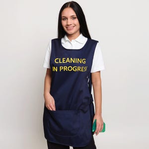 May include: Navy blue apron with a large front pocket and the words "CLEANING IN PROGRESS" in yellow. The apron is worn over a white shirt. A green spray bottle is held in the right hand.