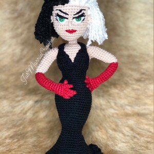 May include: A crocheted doll of Cruella de Vil from Disney's 101 Dalmatians. The doll is wearing a black dress with a plunging neckline and red gloves. Her hair is black and white, and she has green eyes and red lips.