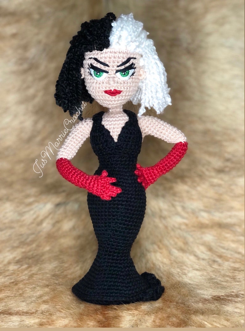 May include: A crocheted doll of Cruella de Vil from Disney's 101 Dalmatians. The doll is wearing a black dress with a plunging neckline and red gloves. Her hair is black and white, and she has green eyes and red lips.