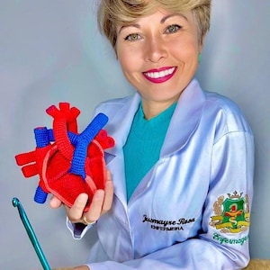 May include: A crocheted anatomical heart model featuring red and blue yarn sections, held alongside a teal crochet hook. The heart displays detailed chambers and vessels, crafted with a textured stitch pattern in vibrant primary colors.