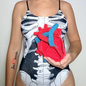 May include: A one-piece swimsuit featuring a black and white anatomical skeleton print with thin black straps. A three-dimensional, red and blue crocheted anatomical heart model is held in front of the torso. The text Jus Marrie Crochet is visible.