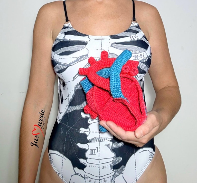 May include: A one-piece swimsuit featuring a black and white anatomical skeleton print with thin black straps. A three-dimensional, red and blue crocheted anatomical heart model is held in front of the torso. The text Jus Marrie Crochet is visible.