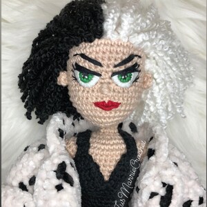 May include: Crocheted doll of Cruella de Vil from the Disney movie 101 Dalmatians. The doll is wearing a black dress and a white fur coat with black spots. The doll has black and white hair, green eyes, and red lips. The doll is holding a red glove in one hand. The doll is made by "Jus Marnie Crochet."