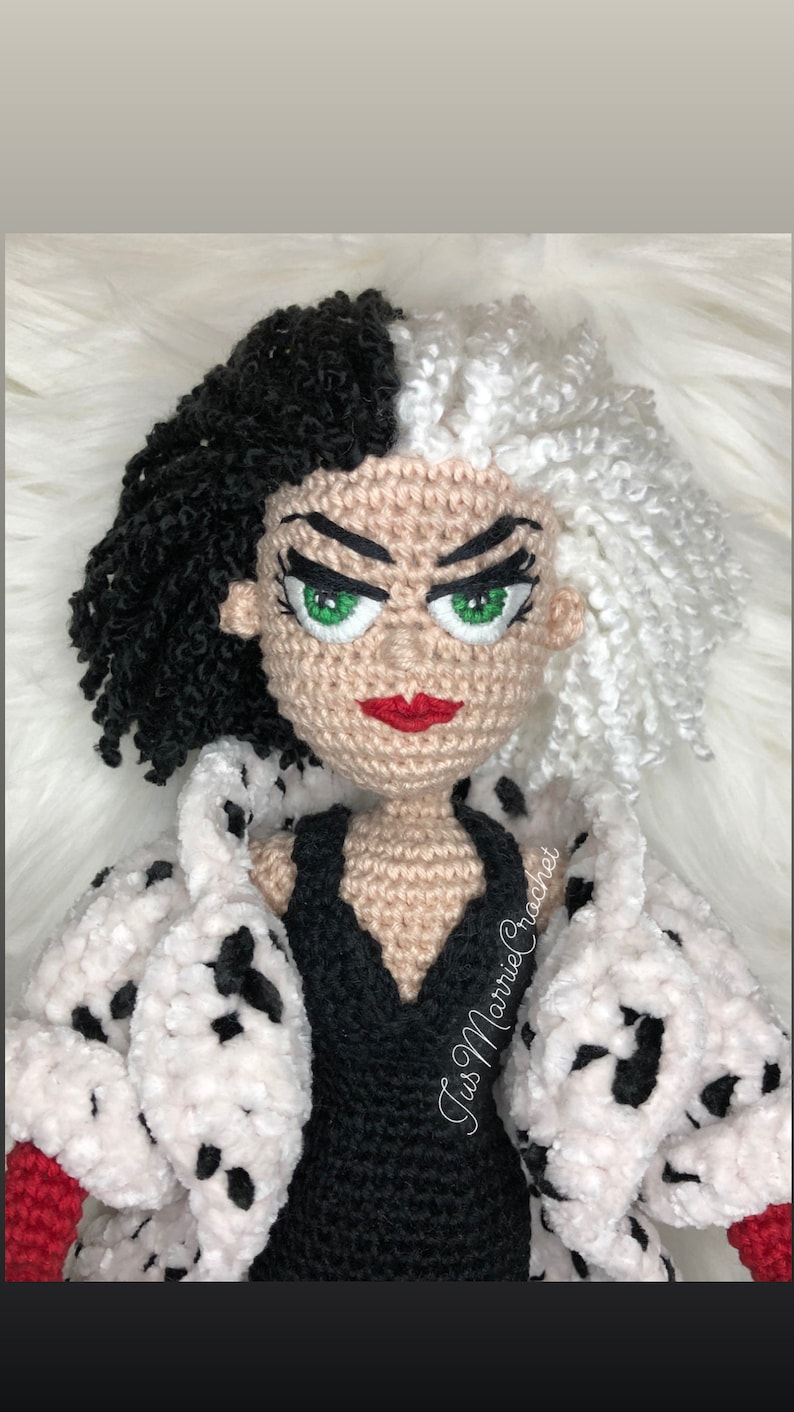 May include: Crocheted doll of Cruella de Vil from the Disney movie 101 Dalmatians. The doll is wearing a black dress and a white fur coat with black spots. The doll has black and white hair, green eyes, and red lips. The doll is holding a red glove in one hand. The doll is made by "Jus Marnie Crochet."