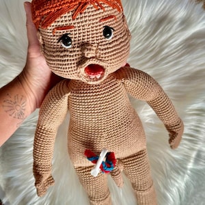 Puede incluir: Una muñeca de crochet con una peluca roja, piel marrón y una cara detallada. La muñeca lleva un atuendo de crochet blanco y tiene un corazón de crochet rojo y azul en el pecho.