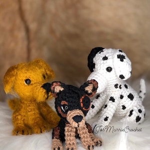 May include: Three crocheted dog toys. One is yellow, one is black and brown, and one is white with black spots. The white dog has a black nose and black eyes. The black and brown dog has a brown nose and black eyes. The yellow dog has a black nose and brown eyes. The toys are sitting on a white fluffy surface. The text "Tiss Marrie Crochet" is visible in the bottom right corner of the image.