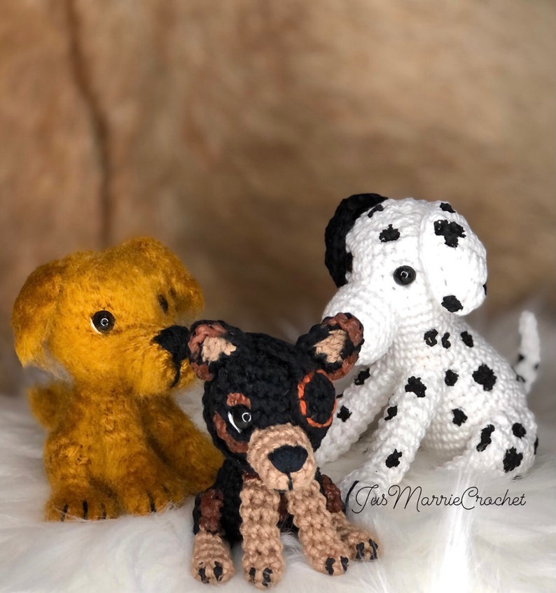 May include: Three crocheted dog toys. One is yellow, one is black and brown, and one is white with black spots. The white dog has a black nose and black eyes. The black and brown dog has a brown nose and black eyes. The yellow dog has a black nose and brown eyes. The toys are sitting on a white fluffy surface. The text "Tiss Marrie Crochet" is visible in the bottom right corner of the image.