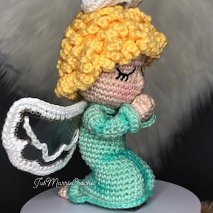 Puede incluir: Una figurita de ángel de crochet con un halo blanco, cabello amarillo y un vestido turquesa. El ángel está arrodillado con las manos juntas en oración.