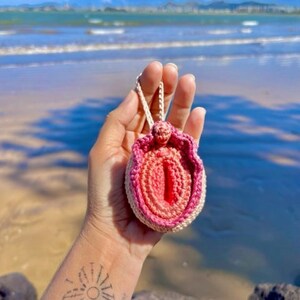 Puede incluir: Un coraz&oacute;n anat&oacute;mico de crochet rosa y blanco, con una cuerda blanca atada, sostenido en una mano contra un fondo de playa.