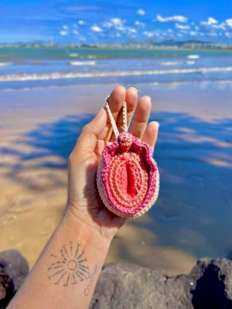 Puede incluir: Un coraz&oacute;n anat&oacute;mico de crochet rosa y blanco, con una cuerda blanca atada, sostenido en una mano contra un fondo de playa.