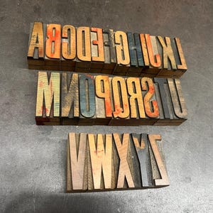 May include: Three sets of vintage wooden letterpress printing blocks. The blocks are arranged in rows, displaying uppercase letters in various colors, including orange, blue, and brown. The blocks are on a gray surface.