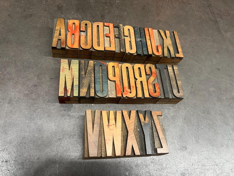 May include: Three sets of vintage wooden letterpress printing blocks. The blocks are arranged in rows, displaying uppercase letters in various colors, including orange, blue, and brown. The blocks are on a gray surface.