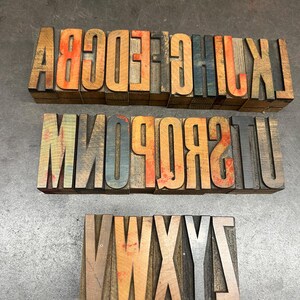 May include: Three sets of vintage wooden letterpress printing blocks. The letters are in various colors, including orange, blue, and brown. The blocks are arranged in rows, with each row displaying a different set of letters.