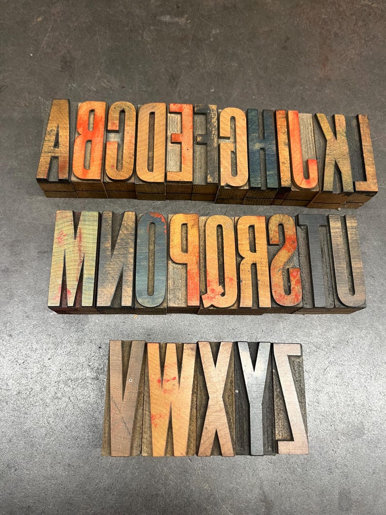 May include: Three sets of vintage wooden letterpress printing blocks. The letters are in various colors, including orange, blue, and brown. The blocks are arranged in rows, with each row displaying a different set of letters.