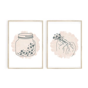 May include: Two framed art prints. One features a jar with flowers, the other shows a hand holding flowers. Both have a light pink watercolor background and dark teal line art. The frames are light wood.