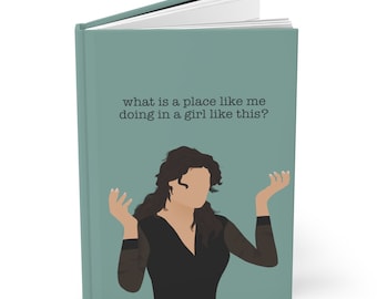May include: A teal journal with the text "what is a place like me doing in a girl like this?" and an illustration of a woman with her arms raised. The journal has a hard cover and white pages.