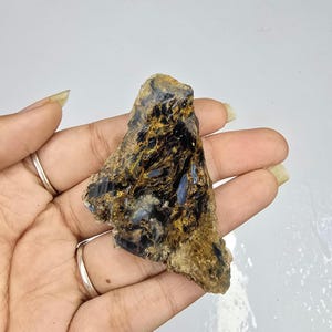 Puede incluir: Una gema en bruto con una mezcla de colores, incluyendo negro, dorado y azul. La piedra tiene una apariencia en capas y se sostiene en una mano, mostrando su textura natural y patrones únicos. Mineral natural.