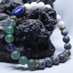 Puede incluir: Una pulsera de cuentas con una variedad de piedras preciosas. La pulsera incluye cuentas verdes, moradas, blancas y grises, junto con una cuenta azul cilíndrica. Espaciadores dorados con cristales transparentes separan algunas de las cuentas. La pulsera se muestra sobre un fondo oscuro y texturizado.