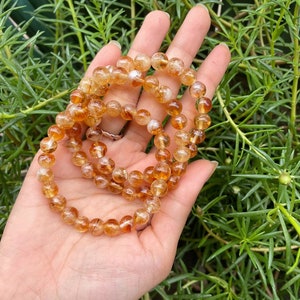 May include: A collection of golden-orange gemstone bead bracelets. The beads are round and translucent, with variations in color. The bracelets are displayed on a hand, with a backdrop of green foliage. The bracelets appear to be made of citrine.