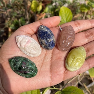 May include: A collection of five carved gemstone pendants, each depicting a serene Buddha face. The stones vary in color, including white, blue, pink, green, and yellow. The pendants are held in an open hand, showcasing their intricate details.