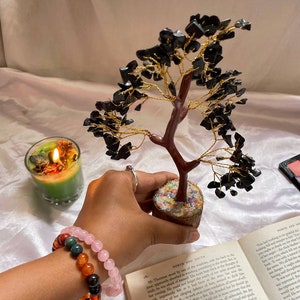Puede incluir: Un árbol de alambre envuelto en piedras preciosas negras con una base de madera marrón. El árbol está sentado en un libro con el texto "NORTH AND SOUTH" visible. Una mano sostiene un brazalete de cuentas de color rosa y multicolor.