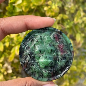 May include: A round, green and black gemstone carved with a lion's face. The gemstone has a polished surface and features red inclusions. The lion's mane and facial features are intricately detailed.