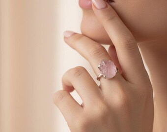 May include: A silver ring featuring a large, irregular-shaped, light pink gemstone. The ring is set on a delicate silver band. The gemstone has a rough, natural texture, and the ring is displayed on a hand.