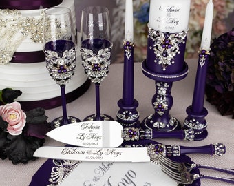 May include: Purple wedding cake knife and server set with a white handle and rhinestones. The set is on a purple plate with the words "Mr. & Mrs. Moyo 02/26/2023" engraved on it. Two purple candlesticks with white candles and a purple candle holder with a white candle are in the background.