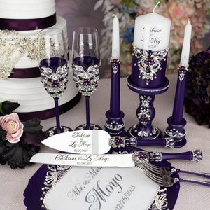 May include: Purple wedding cake knife and server set with a white handle and rhinestones. The set is on a purple plate with the words "Mr. & Mrs. Moyo 02/26/2023" engraved on it. Two purple candlesticks with white candles and a purple candle holder with a white candle are in the background.