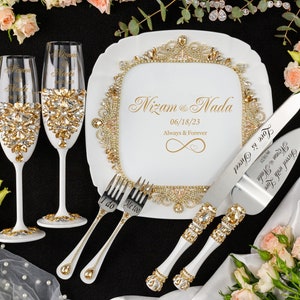 May include: White wedding cake serving set with gold accents. The cake knife and server have rhinestones and the words "Served with Love" and "Love is Sweet" engraved on them. The cake plate has a gold border with rhinestones and the words "Nizam & Nada 06/18/23 Always & Forever". Two champagne flutes with gold and rhinestone accents are also included.