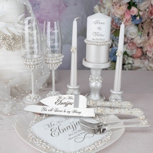 May include: Wedding cake cutting set with a white cake knife and fork, a white cake plate with pearl accents, two champagne flutes with pearl accents, and a white candle with pearl accents. The cake knife and fork are engraved with the text "Mr. & Mrs. Elnajjar You will Forever be my Always". The cake plate is engraved with the text "Mr. & Mrs. Elnajjar 06/10/202 Always & For". The champagne flutes are engraved with the text "Anna & Victor 06/10/2022". The candle is engraved with the text "Anna & Victor 06/10/2022".