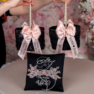 May include: Two black and white sequined ring bearer pillows with pink satin bows and personalized ribbons. The ribbons say "Here comes the bride", "We never fades away", and "Toni Juan July 20 2024". A black satin pillow with a silver embroidered design and the text "July 20 2024 We do!" is in the center.