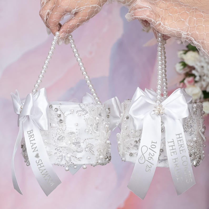 May include: Two white flower girl baskets with pearl handles and satin bows. One basket has the text "Brian &hearts; Shawna" on the ribbon and the other has the text "Here Comes The Bride" and "10/26/25" on the ribbon.