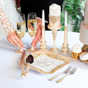 May include: A wedding set featuring champagne flutes, candles, and a serving set. The flutes and serving set have gold and crystal detailing. The candles are cream and white with gold accents. The serving plate has the text "Mr. & Mrs. Tirk 10.11.2020".