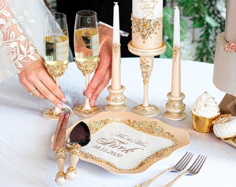 May include: Wedding champagne flutes, candles, and serving set. The flutes and serving set are adorned with gold and crystal accents. The candles are white and cream with gold embellishments. The serving plate has the text "Mr. & Mrs. Tirk 10.11.2020".