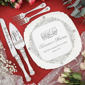 May include: A white cake server and knife set with silver handles and rhinestones. The server has the text "Mr. & Mrs. Faheem 03/10/2023 You will Forever be my Always" engraved on it. The knife has the text "I do" engraved on it. The set is on a white cake plate with a silver border and rhinestones. The plate has the text "Kareem & Mariam And We Created You In Pairs (Quran 78:8)" engraved on it.