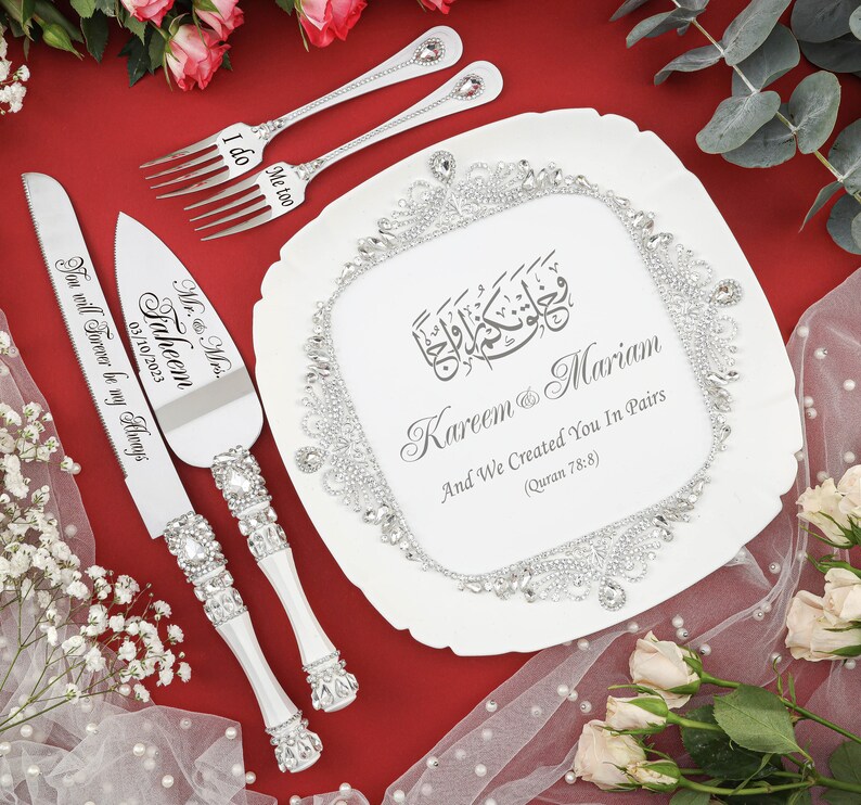 May include: A white cake server and knife set with silver handles and rhinestones. The server has the text "Mr. & Mrs. Faheem 03/10/2023 You will Forever be my Always" engraved on it. The knife has the text "I do" engraved on it. The set is on a white cake plate with a silver border and rhinestones. The plate has the text "Kareem & Mariam And We Created You In Pairs (Quran 78:8)" engraved on it.
