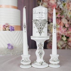 May include: White wedding unity candles with a silver heart and rhinestones. The center candle has the Arabic text "حُبٌّ خَالِدٌ" and the date "08/19/2024".