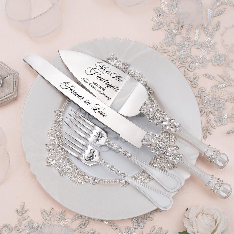 May include: A silver cake knife and server set with two matching forks, featuring handles encrusted with clear crystals. The knife is engraved with "Forever in Love" and the server with "Mr. & Mrs. Pauligoti 08.01.2026 Two hearts, one journey".